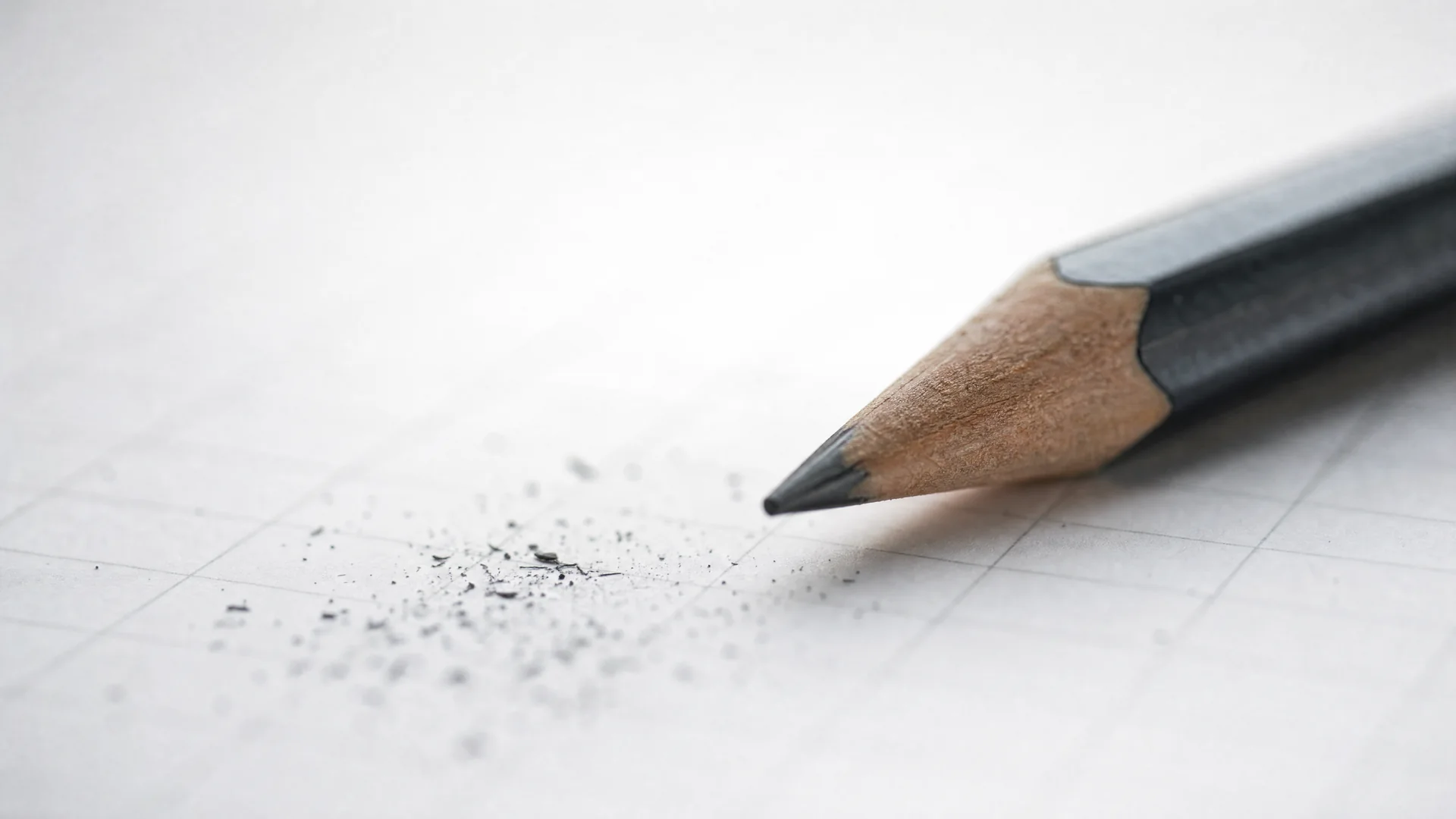A sharpened graphite pencil resting on squared paper at a shallow angle, the grid falling out of focus.